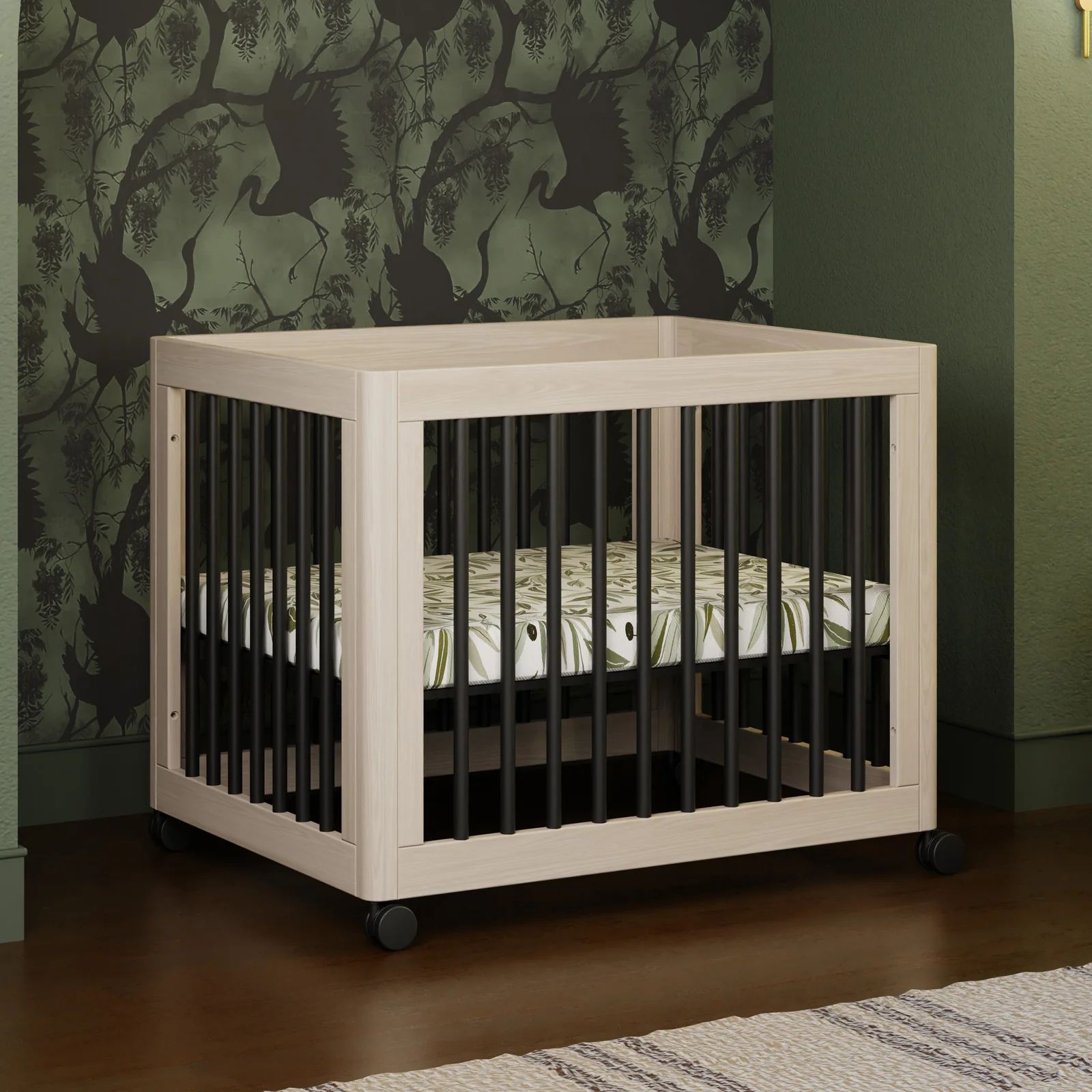Babyletto Yuzu 8-in-1 convertible crib in natural wood with black slats and caster wheels, styled in a modern nursery with dark green crane-pattern wallpaper and a botanical mattress
