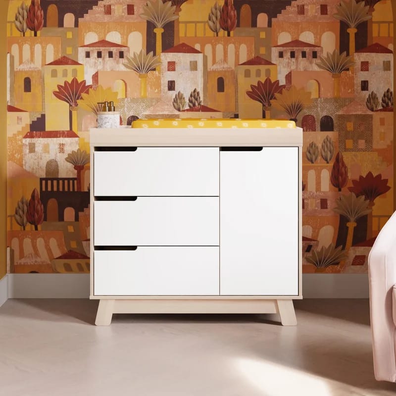 White nursery dresser with storage drawers against a warm patterned wallpaper.