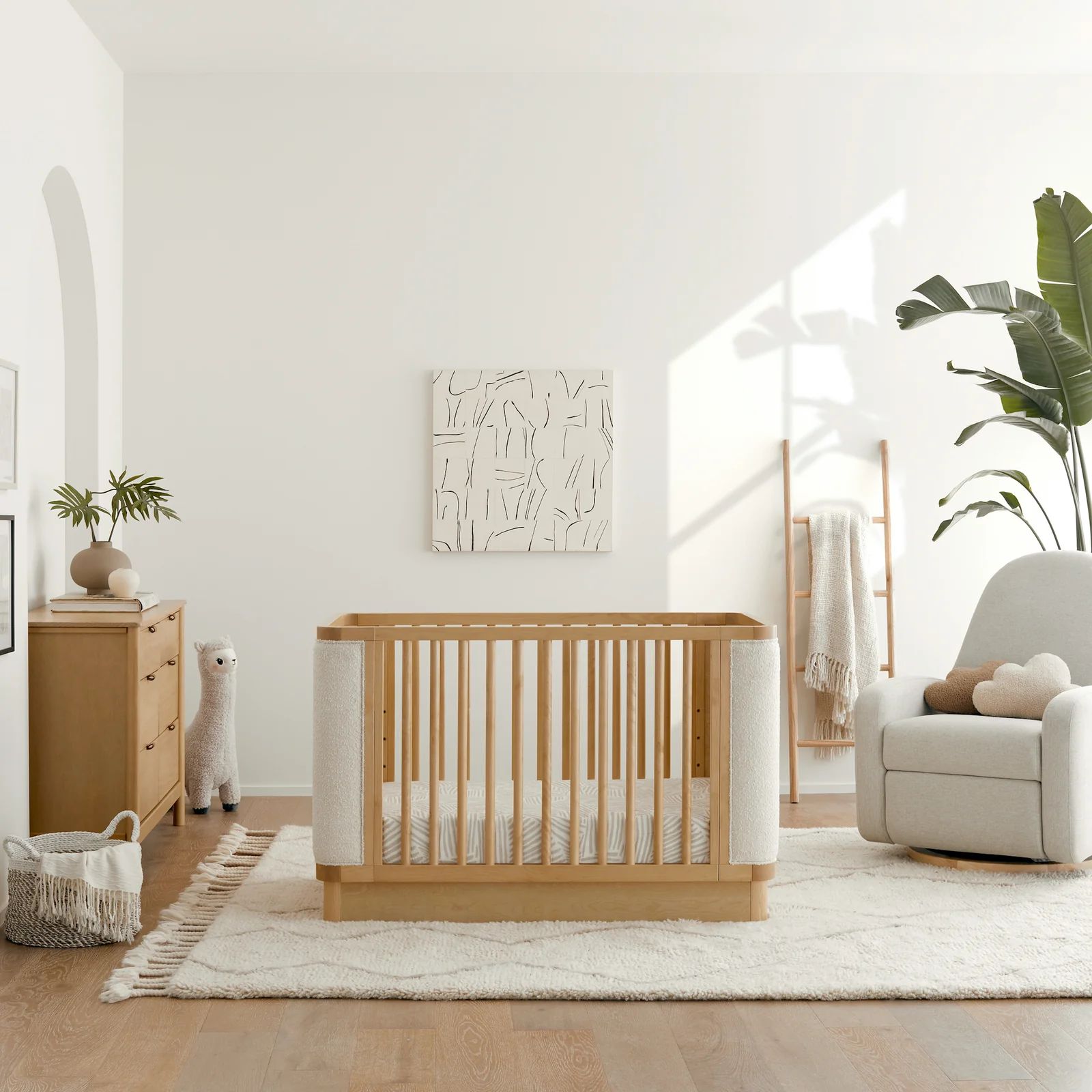 Minimalist nursery featuring a natural wood crib with soft boucle side panels, a cream area rug, wooden dresser, cozy glider chair, large indoor plant, and warm sunlight streaming across white walls