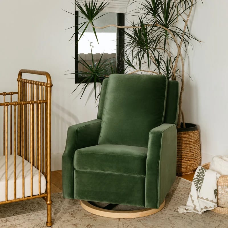 Green velvet nursery rocking chair beside a wooden crib and indoor plants.
