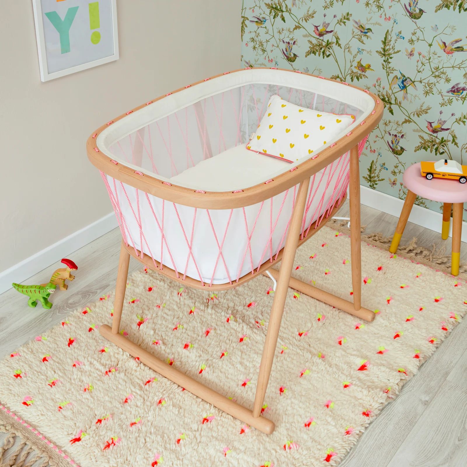 Kumi Bassinet with natural wood frame and pink woven rope sides in a bright nursery with floral wallpaper