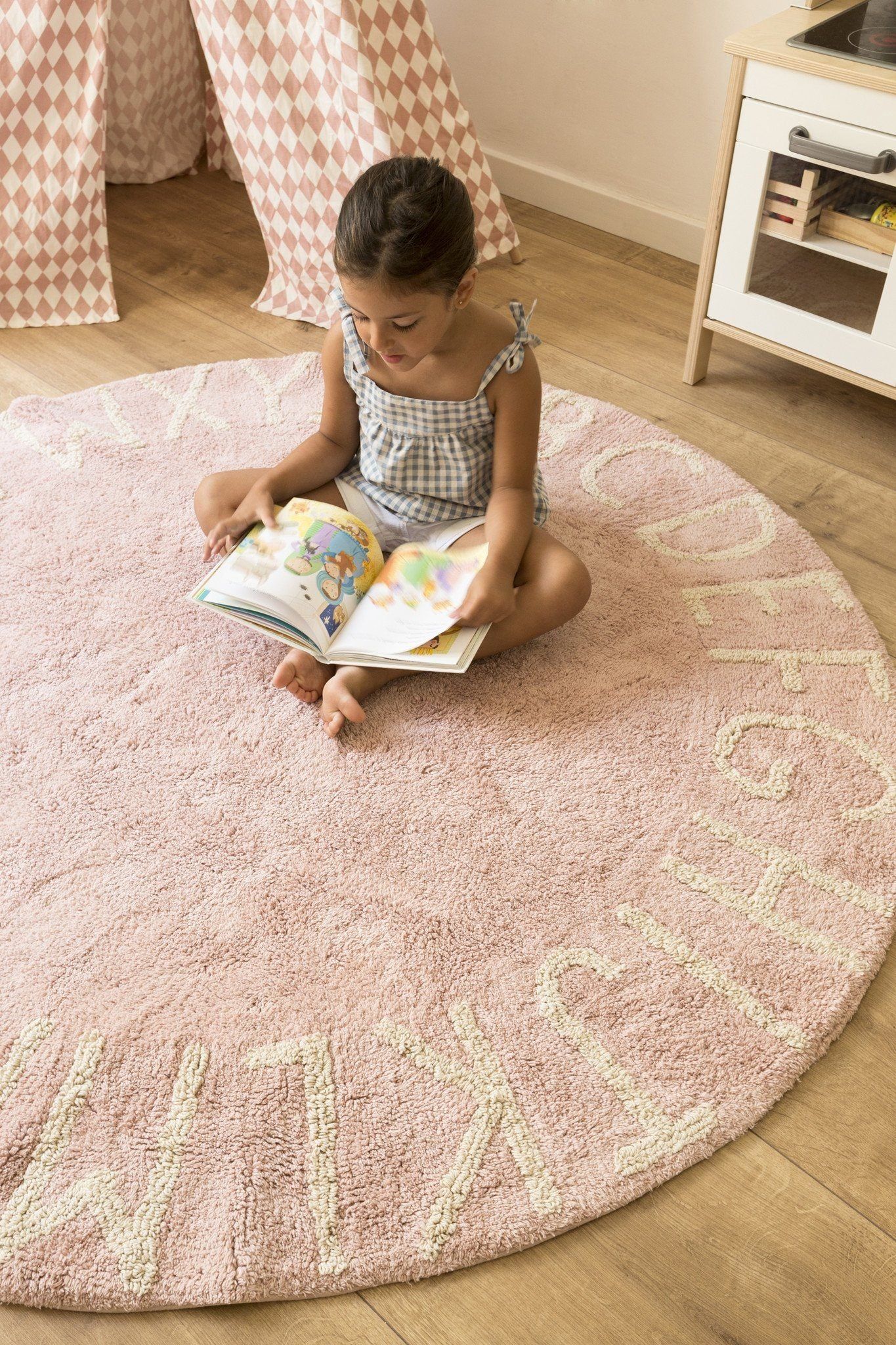 A young girl reading a book while sitting on a soft pink kids’ rug in a cozy playroom.
