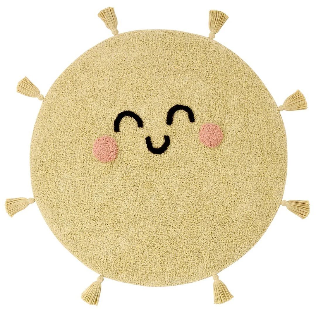 Round yellow kids’ rug with a smiling face design, pink cheeks, and decorative tassels around the edges.
