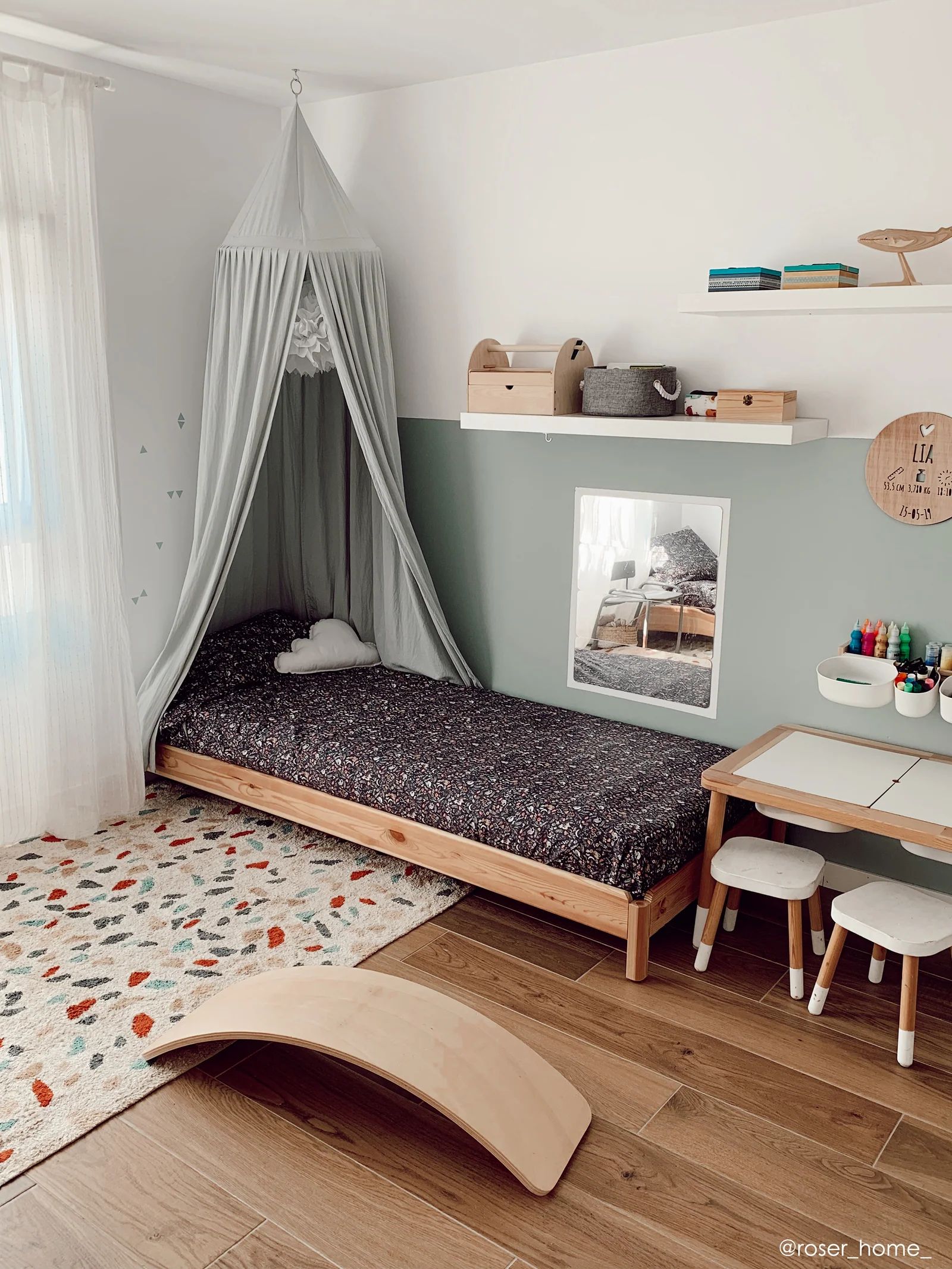 Cozy children’s bedroom with a colorful patterned rug, wooden bed, canopy, play table, and shelves.
